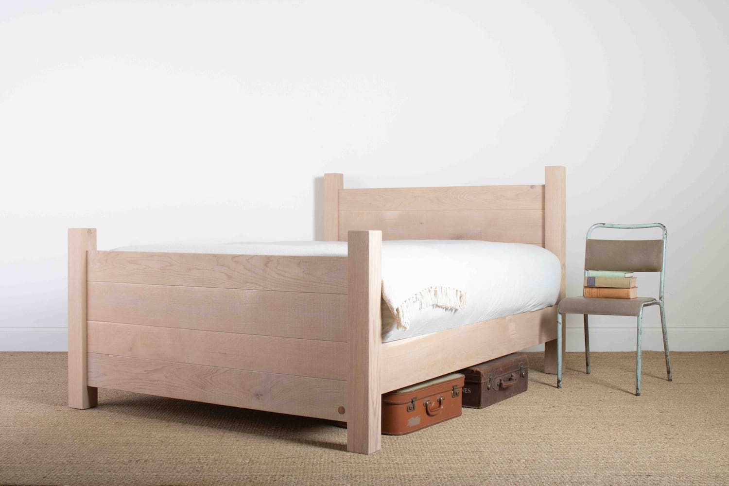 A kingsize oak bed with footboard and chunky square posts sits on a wheatgrass carpet with a plain white wall in the background a metal chair sits beside the oak bed and 2 leather suitcases sit under the bed