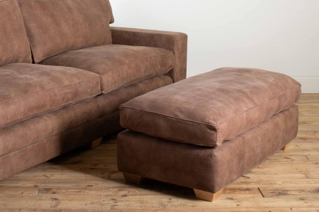 A picture of a large handmade brown leather footstool that has a cushion top featuring piped edges. The low fee are tapered square and modern made from natural coloured wood.