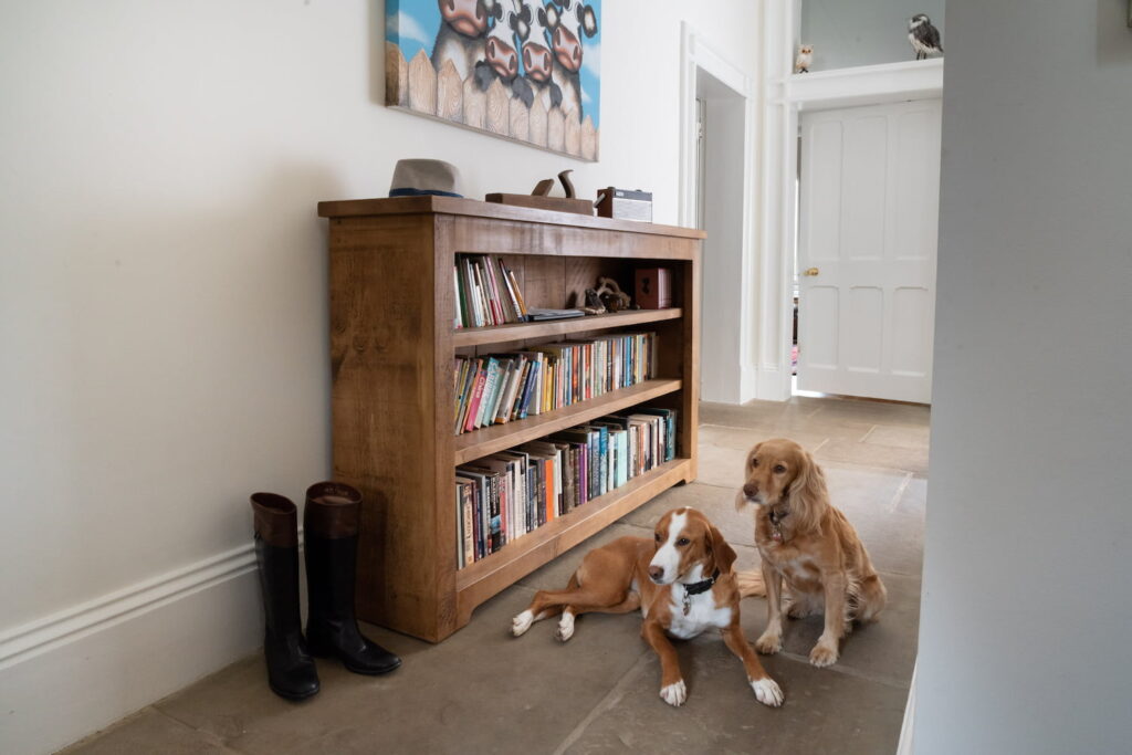 A rustic pine bookcase filled with books in a hallway with natural stone floor with 2 dogs sat beside the wooden bookcase