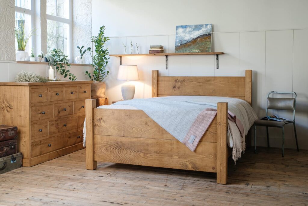 A handmade plank bed sits on a wooden floor in a converted mill with windows letting light in a large chest of solid wood drawers sit beneath the window with plants and artwork displayed around the room