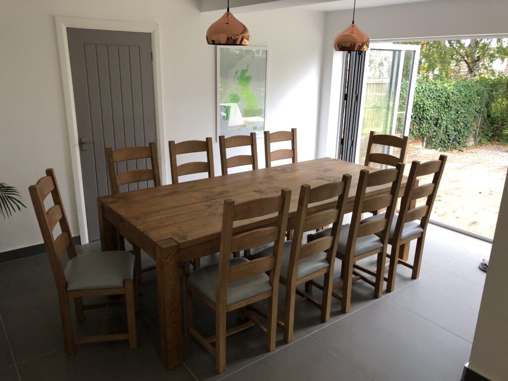 An extra large wooden dining table with 10 chairs sits in a modern home with french doors leading to a garden