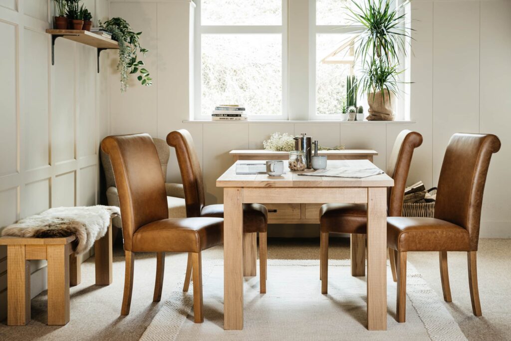 A solid oak farmhouse dining table sits in front of a large window with tall plants on the window cill. The oak dining table has 4 leather chairs beside it with a bench against the wall with a sheepskin on the bench.