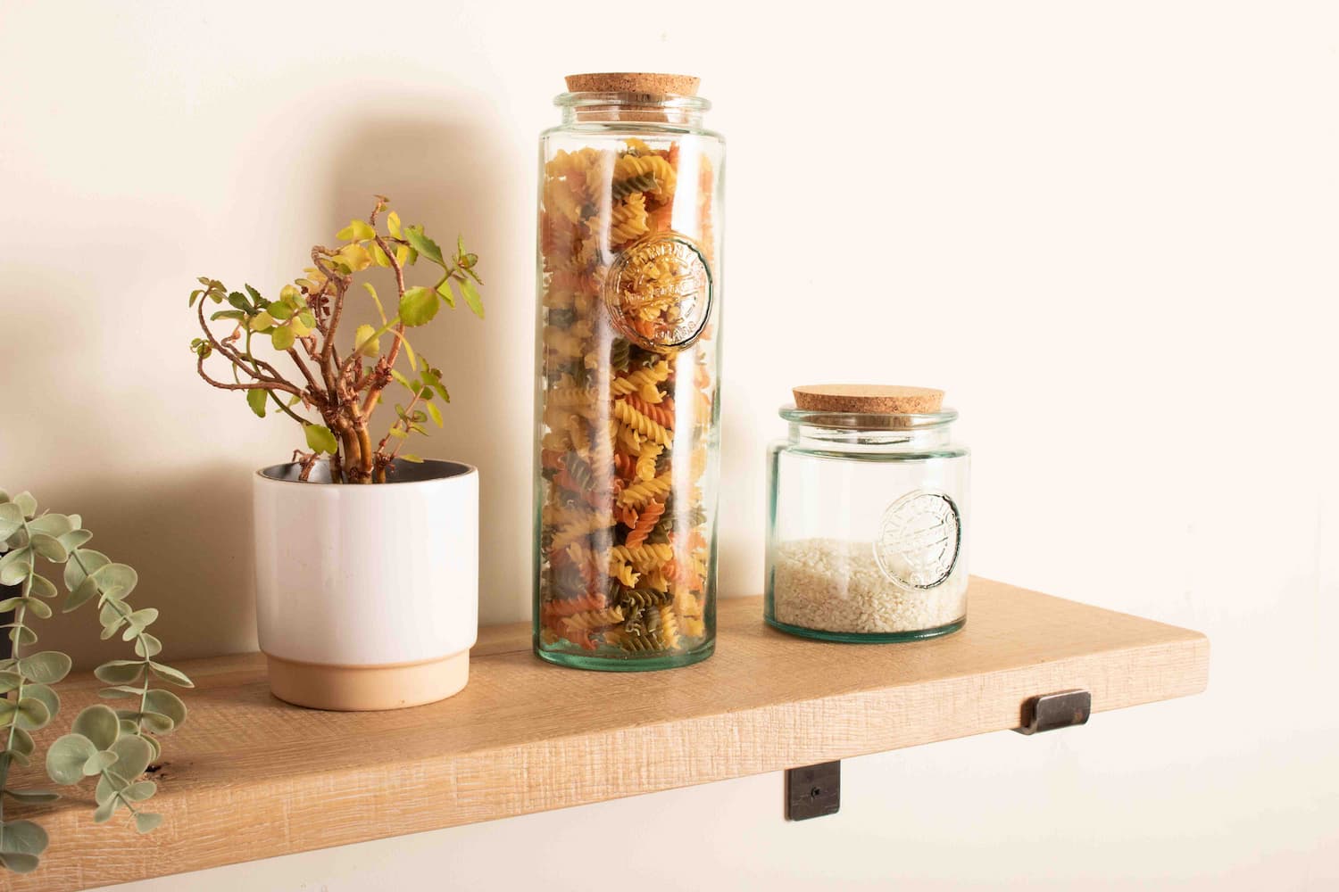 a natural oak shelf is fitted to a white wall with metal shelf brackets. A display of kitchen goods and pot plants is on the wooden shelf