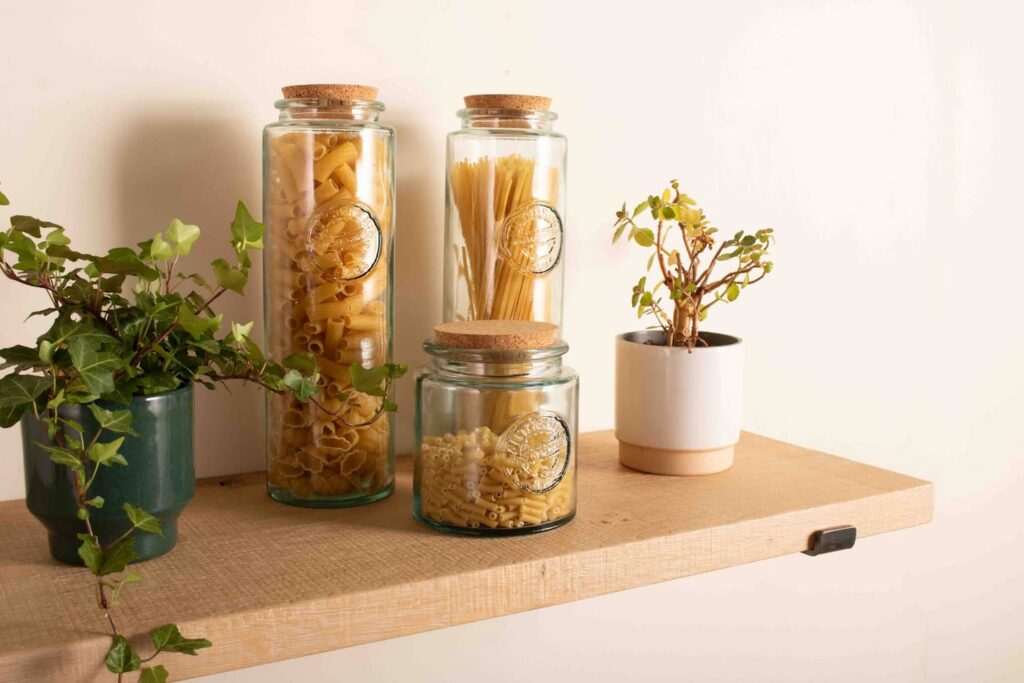 3 glass kitchen storage jars and 2 pot plants sit on a rustic chunky oak shelf thats fitted to a white wall with black metal shelf brackets