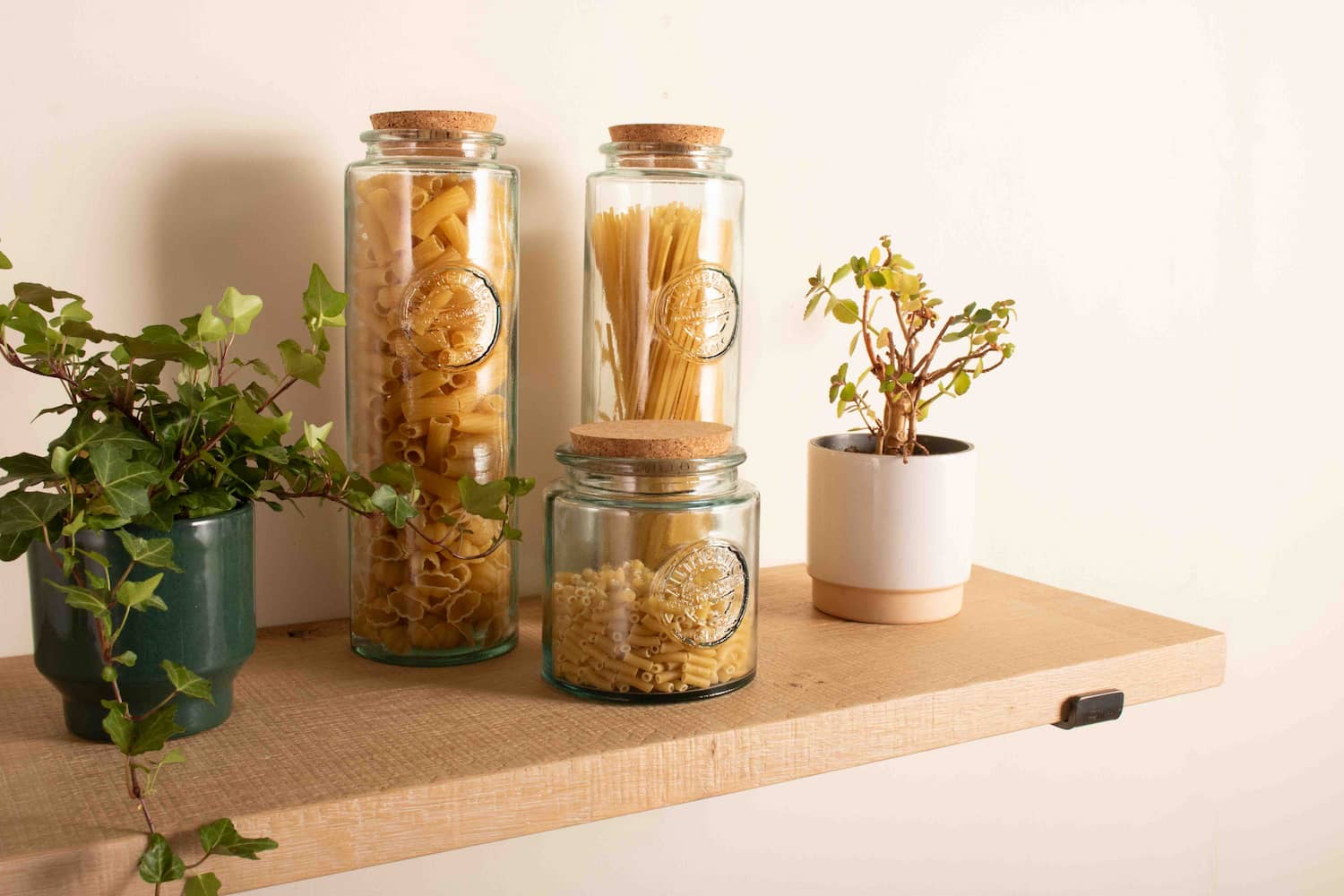 3 glass kitchen storage jars and 2 pot plants sit on a rustic chunky oak shelf thats fitted to a white wall with black metal shelf brackets