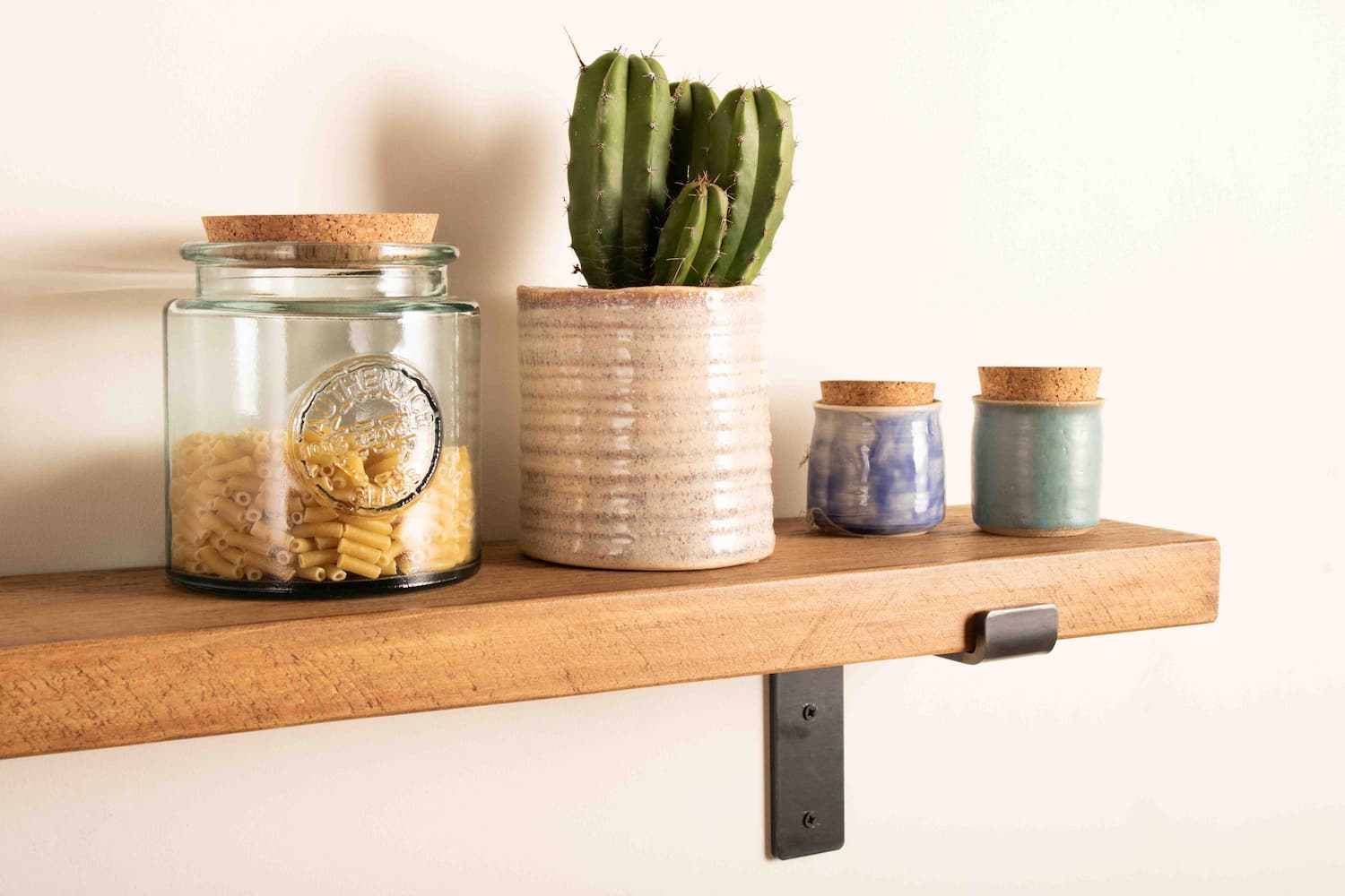 A chunky wooden kitchen shelf is fitted to a white wall with black metal shelf brackets. A display of macaroni in a glass jar and a cork lid sits next to a cactus and little ceramic jars.