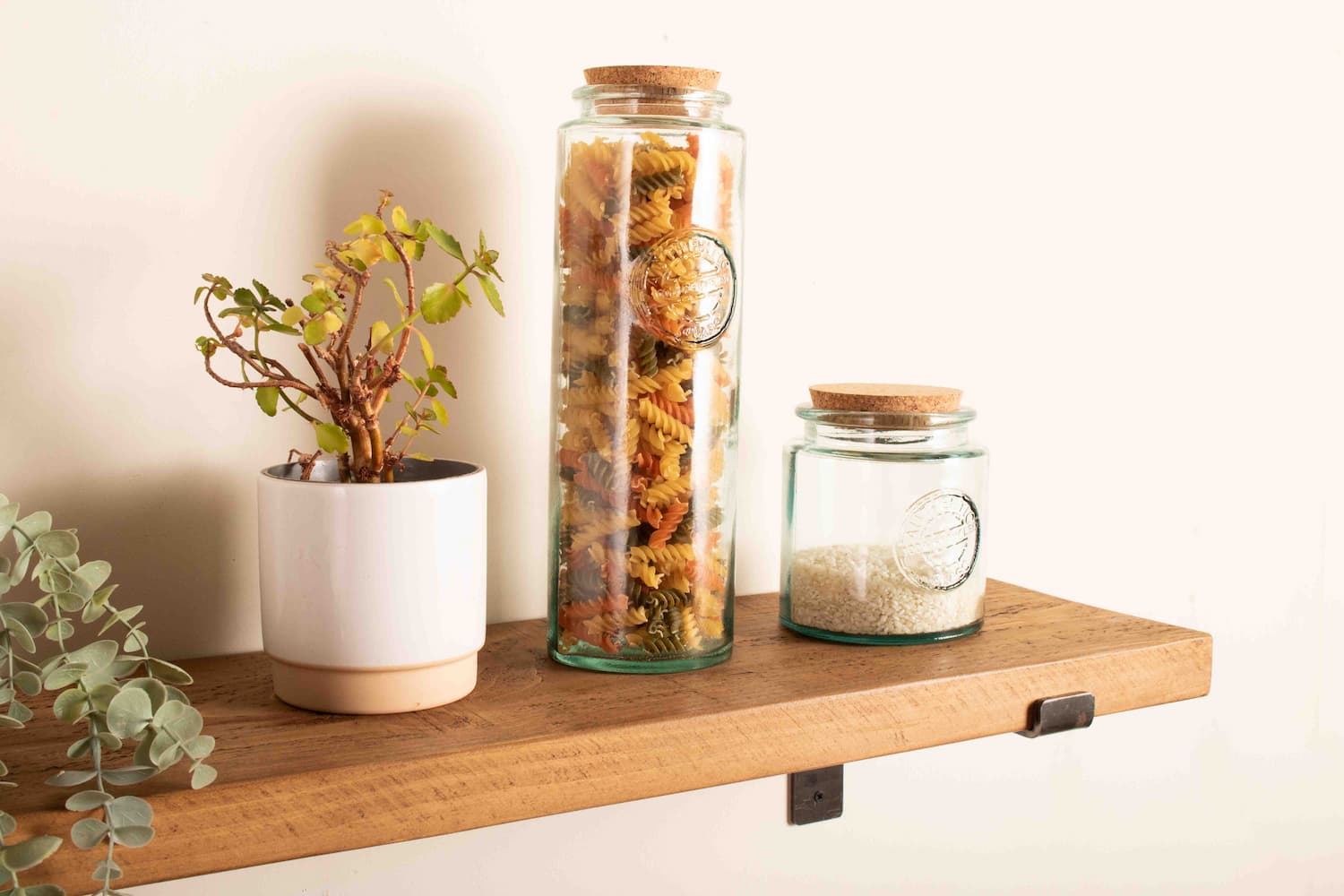 A rustic pine kitchen shelf is featured with a display of pasta and rice in glass jars with cork lids and a plant pot