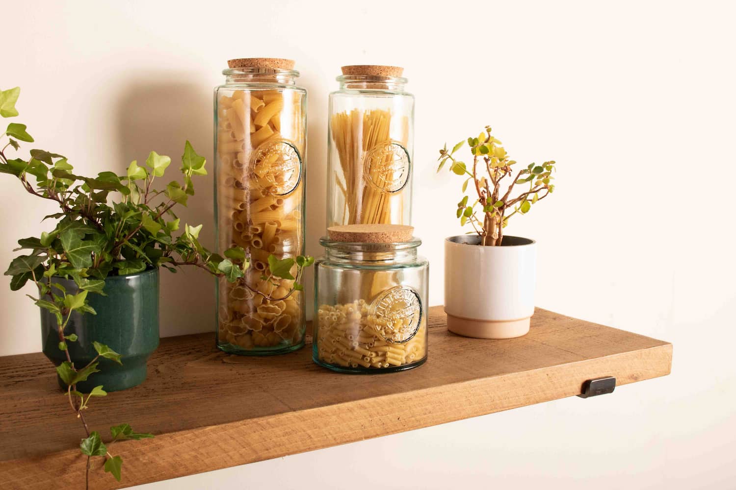 A chunky wooden shelf is shown fitted to a white wall using contemporary steel shelf brackets. On the chunky shelf there is a selection of glass jars with pasta in them and cork lids.