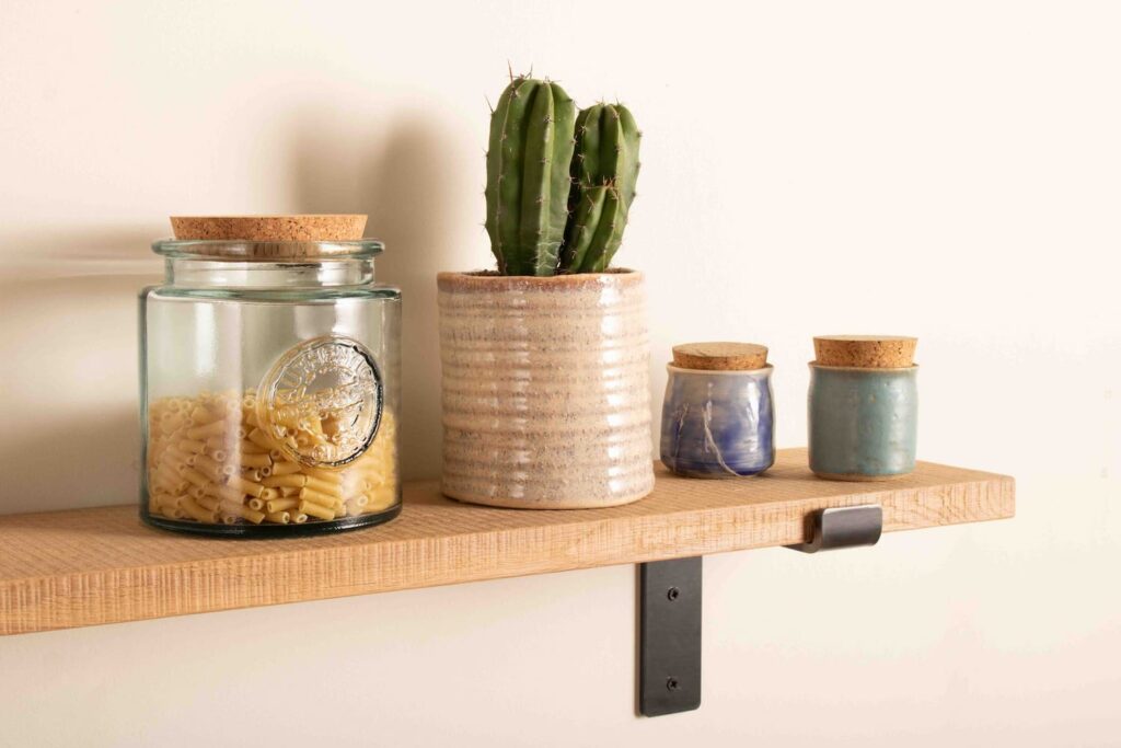 a rustic oak shelf is fitted to a white wall with modern steel shelf brackets. The rustic oak shelf is displaying a cactus in a pot, two small ceramic pots with cork lids and a glass jar with pasta in it and a cork lid