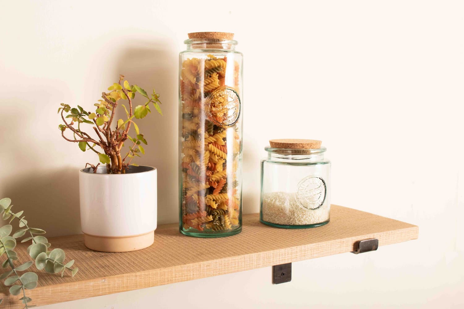 a solid oak shelf is fitted to a plain white wall and the oak shelf is displaying glass jars with pasta and rice in them next to a pot plant