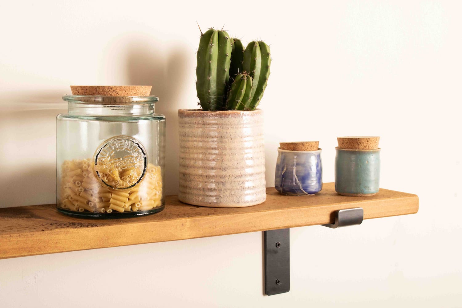 A bespoke wooden shelf is fitted to a wall with metal shelf brackets with a plain white background. the wooden shelving has a cactus in a pot displayed next to small