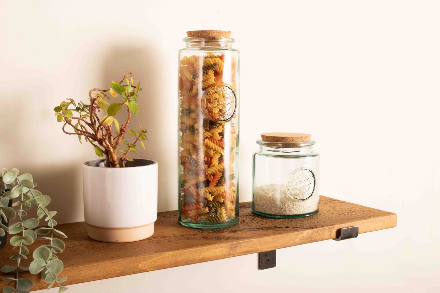 a rustic wooden shelf is fitted to a white wall using metal shelf brackets and glass storage jars with pasta and rice are on display amongst plants in pots