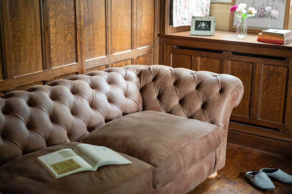 A close up of a brown leather chesterfield sofa beside a small window. The room setting has golden oak panelling on the walls