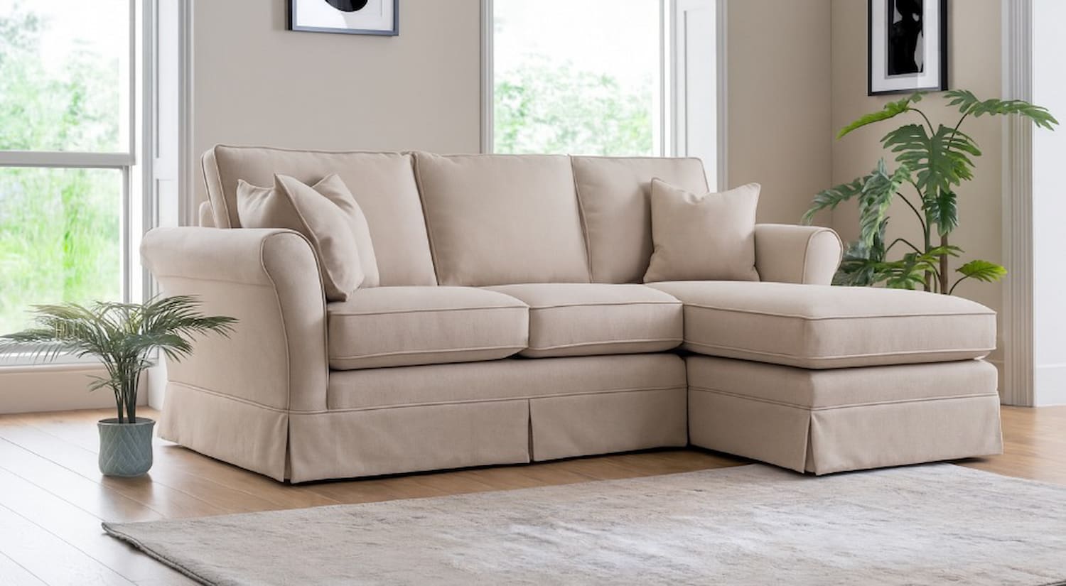 A cream linen loose cover sofa with a chaise extension sits in a modern house with pale walls, modern framed art, plants and a pale rug and large bright windows.